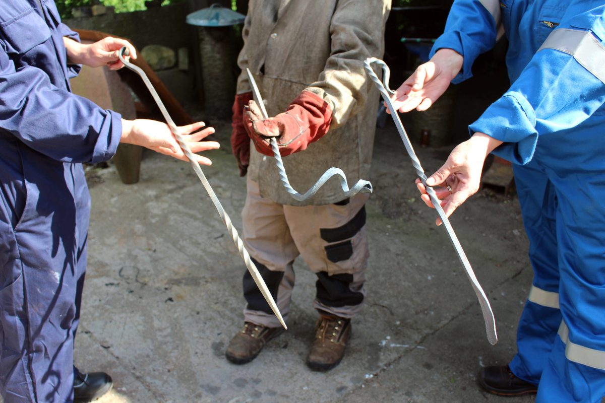 A close-up view of three steel fire pokers being presented in the hands of their respective makers. The left poker has straight, even twists, coming to a point at the end. The centre poker coils off to the maker’s left in a curve. The poker on the right has an ornate looped handle and is flattened at the end.