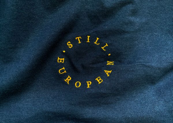 the words 'still european' are embroidered in golden yellow on a navy fabric background