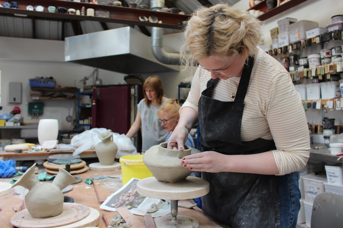A woman with bleached blonde hair wears an apron and leans over a half constructed clay form on a banding wheel in the ceramics studio.