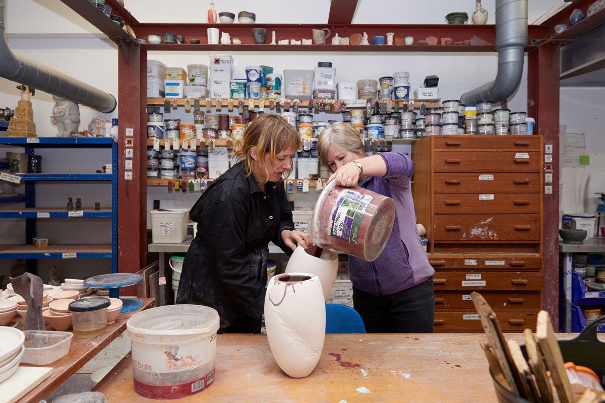 Two people juggle a large ceramic vessel in SSW's Ceramics workshop. One holds the white, bisque-fired vessel and the other pours a brown coloured glaze - from a large tub - into the vessel.