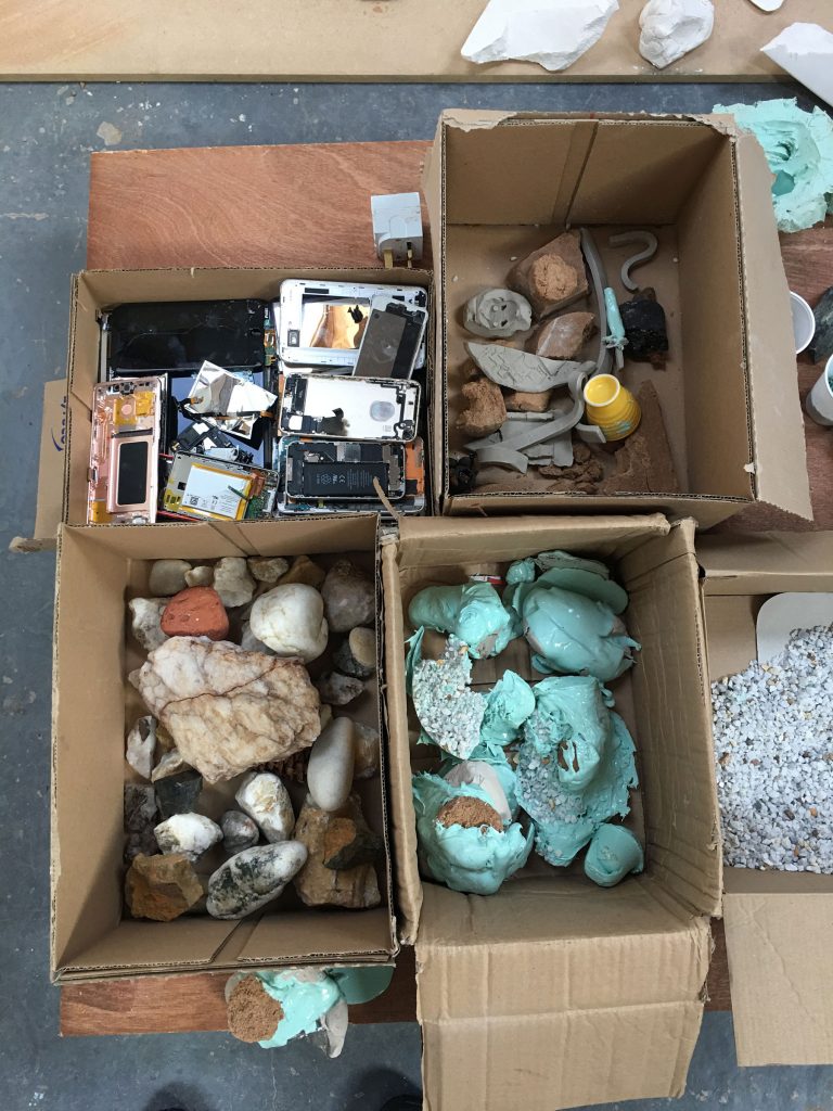 looking down into four cardboard boxes arranged in a grid. The top left contains broken mobile phones, top right bits of clay and rocks, bottom left a selection of rocks and bottom right a selection of casts in turquoise latex. 
