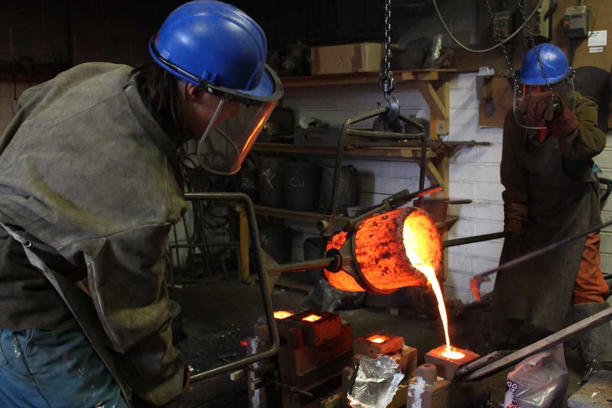 In SSW's foundry, two people wearing blue hardhats, visors and leather jackets use a metal frame suspended from the ceiling to tip a glowing orange crucible of molten bronze towards the camera. The bronze pours into a mould below. 