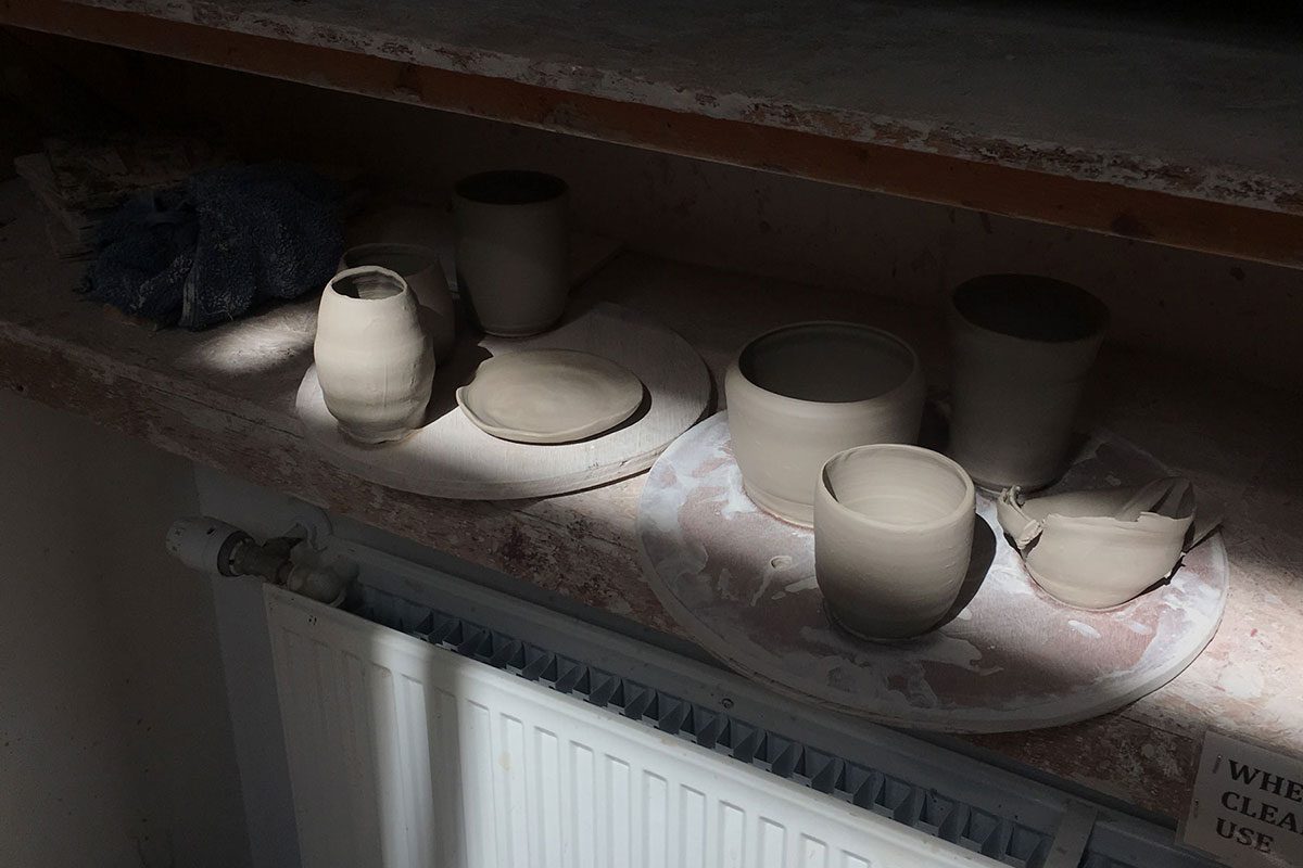 The image is largely cast in shadow, with a dramatic beam of light cutting diagonally across it. The light falls on 2 MDF throwing batts, next to each other on a shelf above a long, white radiator. The batts have some clay vessels on them which vary in form and are a light, sandy grey in colour.