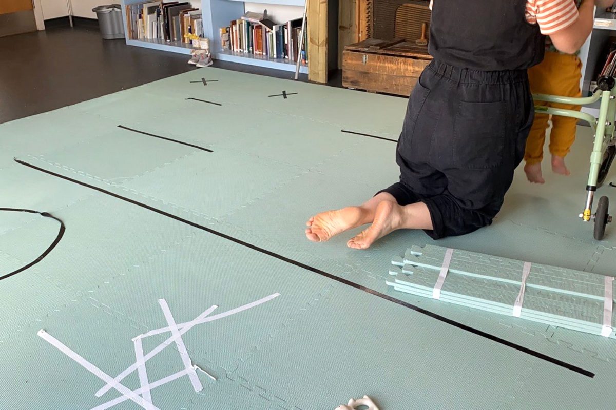 Duck egg blue mats cover a lino floor. They have white and black taped symbols all over. In the top right corner of the photo a person wearing a black jumpsuit kneels, facing away from the camera. They have bare feet. They are lifting a child in mustard yellow trousers up from a greeny-blue walking aid.
