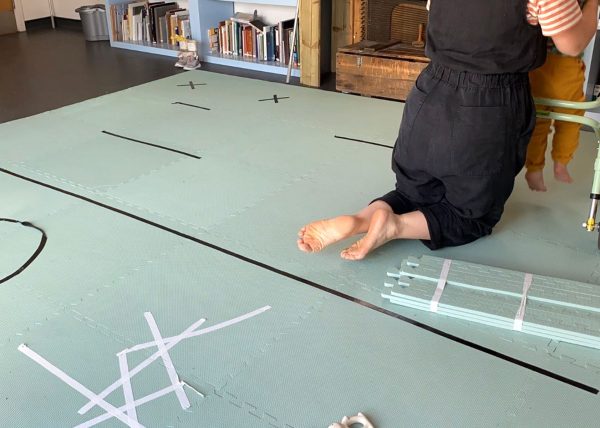 Duck egg blue mats cover a lino floor. They have white and black taped symbols all over. In the top right corner of the photo a person wearing a black jumpsuit kneels, facing away from the camera. They have bare feet. They are lifting a child in mustard yellow trousers up from a greeny-blue walking aid.