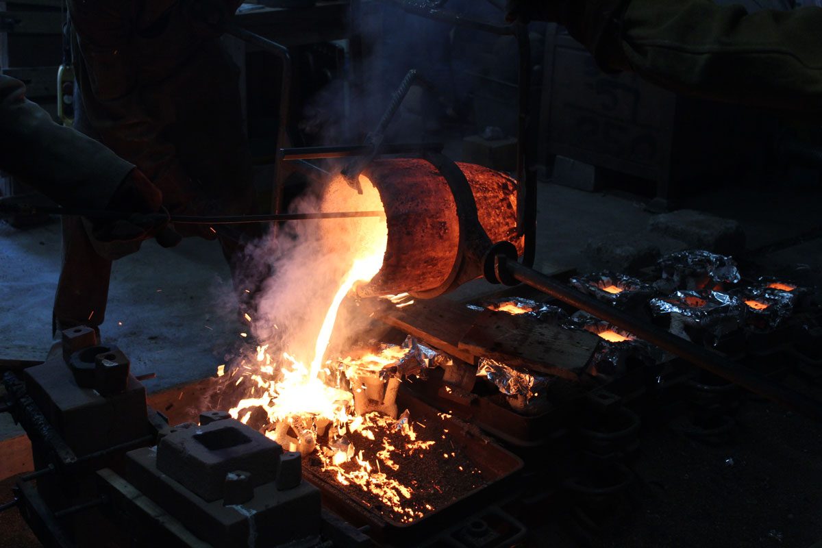 Glowing molten bronze pours out of a crucible into tin-foil covered sand moulds. The metal steams and splashes.