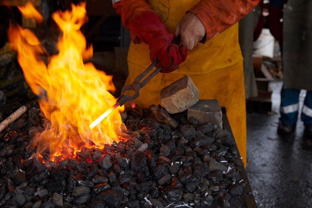A pair of white hands hold a metal bar in a large flame, with tongs. The metal bar glows white with the heat.