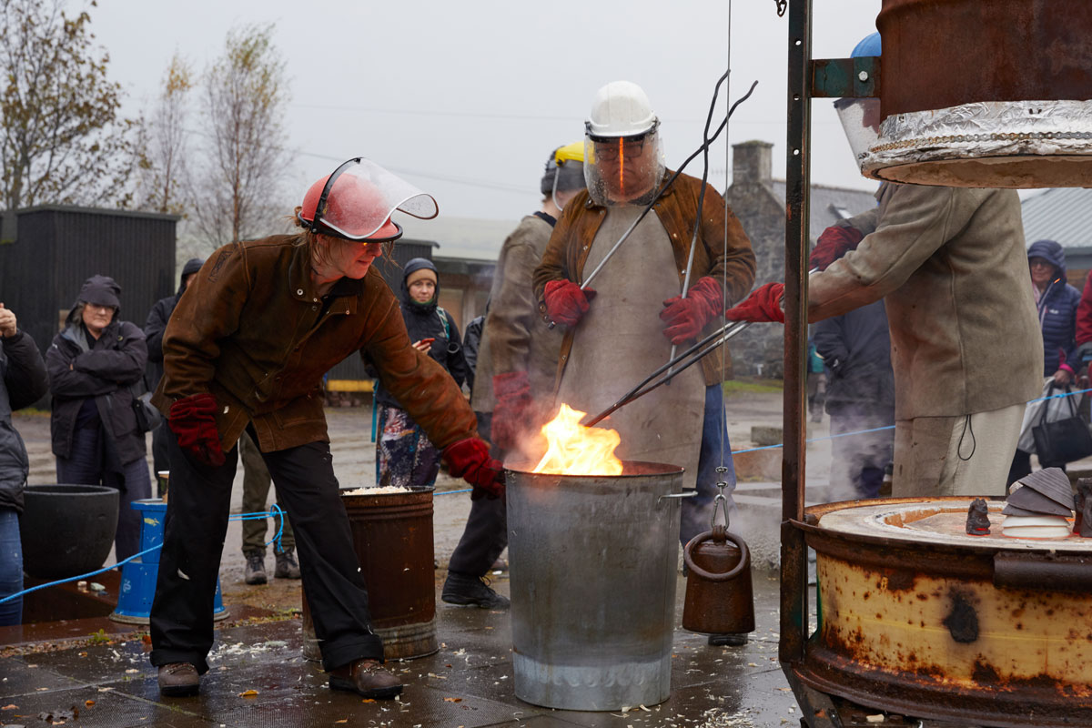 Three people wearing leather jackets and aprons, visors and gloves, lower ceramics into a flaming bin behind a glowing orange raku kiln. A crowd are watching. 
