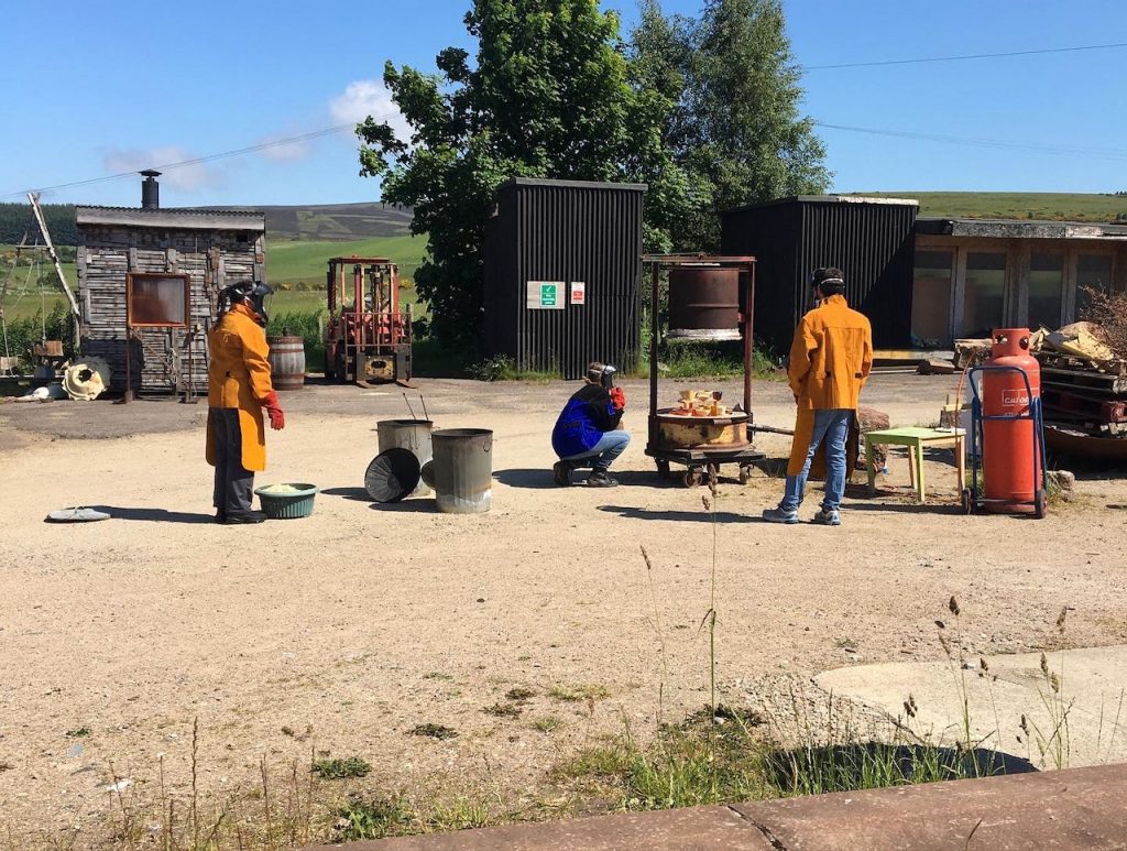 From a distance, Beth the ceramics technician wearing a deep cobalt leather jacket and visor crouches to look at the open contents of the raku kiln. Two other people wearing orange leathers and visors stand nearby. Raku kit such as metal dustbins and a large canister of propane are dotted around. They are in the SSW yard and it is gloriously sunny. 