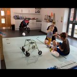 Laura and Murray sit on light blue foam mats on the floor and hold Rowan and Jude's hands. Rowan and Jude are leaning on Laura and Murray for support to stand up. Rowans waking frame is in the foreground and buggy is in the background.