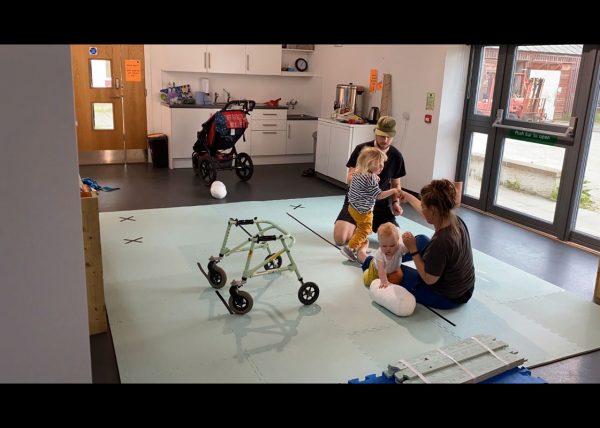 Laura and Murray sit on light blue foam mats on the floor and hold Rowan and Jude's hands. Rowan and Jude are leaning on Laura and Murray for support to stand up. Rowans waking frame is in the foreground and buggy is in the background.