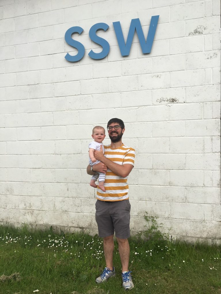 A white man with dark hair, glassws and a beard holds a young baby with blonde hair and smiles at the camera. He is wearing a stripey yellow t-shirt and shorts, and stands on mowed grass in front of a whitewashed wall. The letters SSW are in blue above his head. 