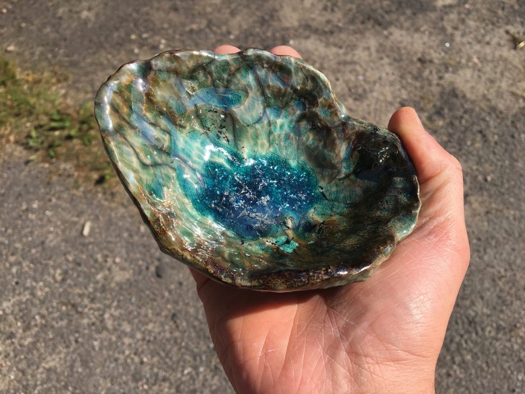 A wobbly ceramic bowl held in a person's hand. It looks like a crystal with browns and blues sparkling in the sunshine. The centre is deep blue and sparkles enticingly. 