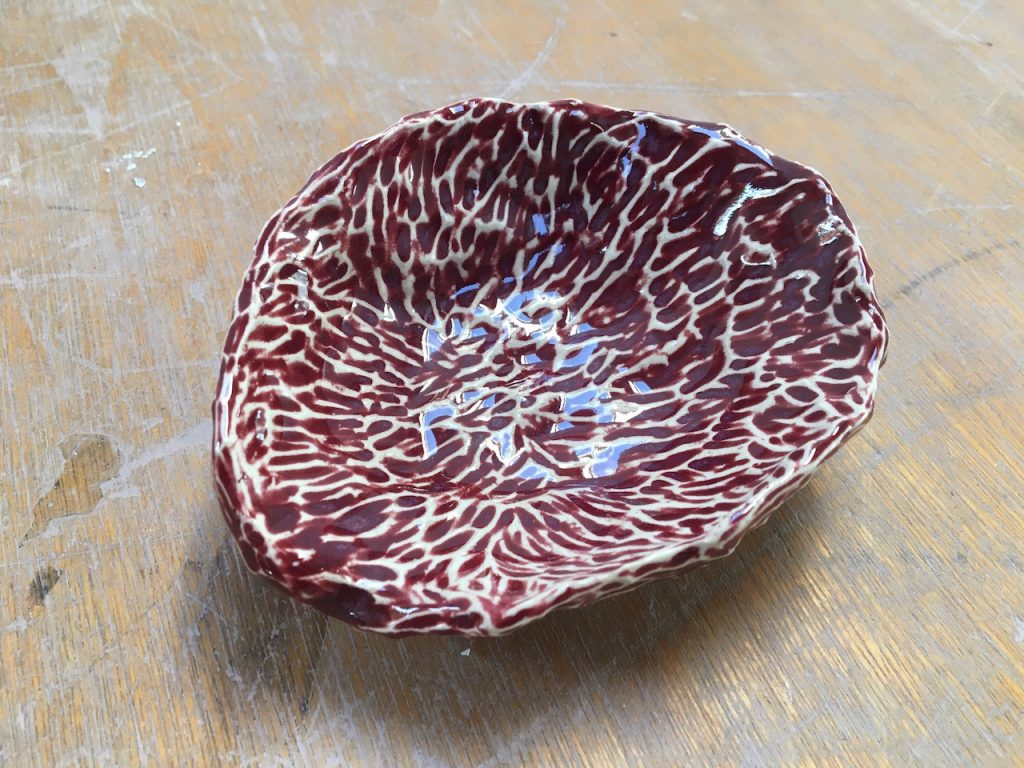 A wobbly ceramics bowl. It is coloured deep purple with white 'veins' running all over. The veins look like they protrude from the surface. It is shiny and looks small as it sits on a wooden surface where you can see the grain. 