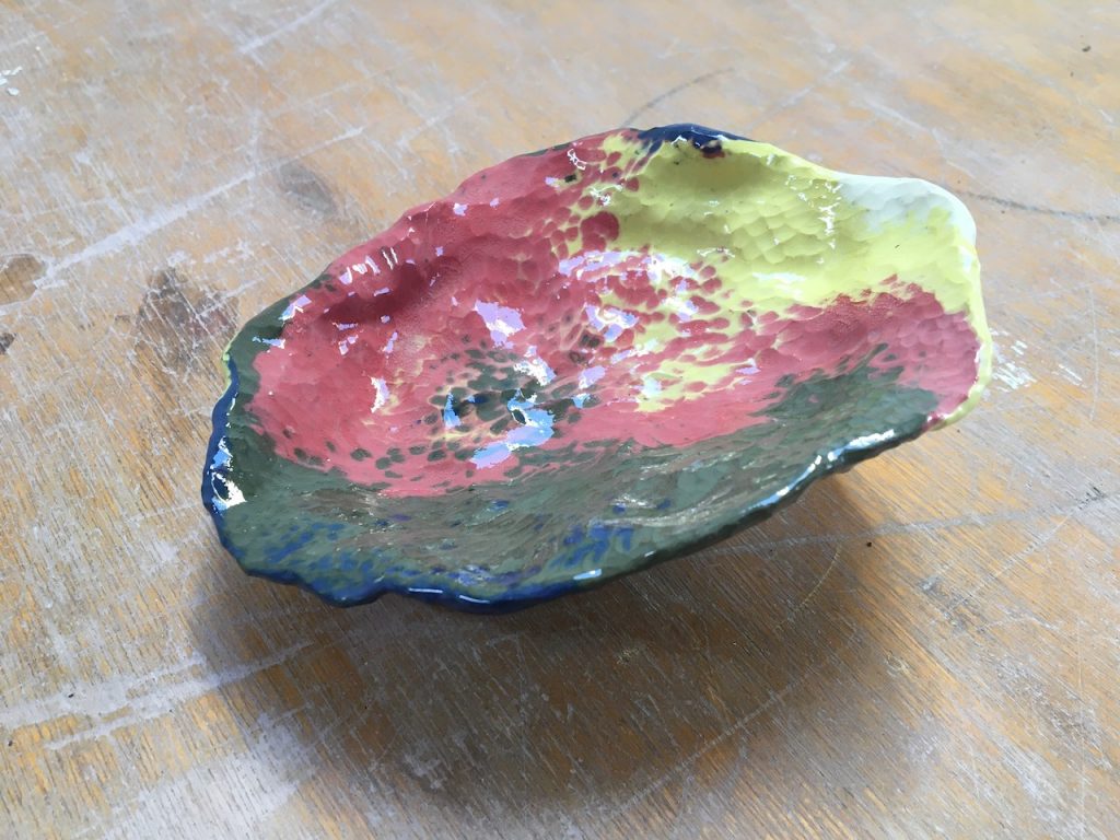 A close up of a wobbly, oval shaped ceramic form, coloured pink, yellow and green. The surface is shiny and bumpy. It looks quite small as it is sitting on a wooden surface and the grain is visible. 