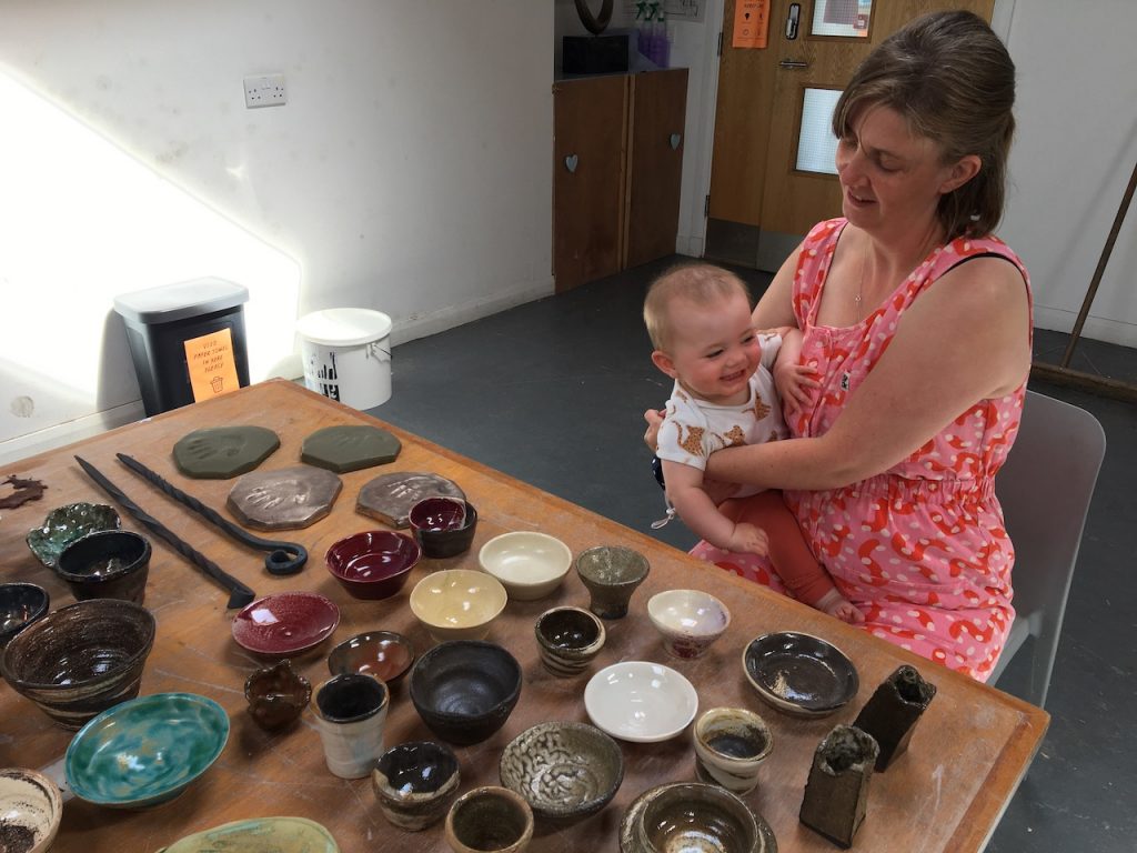 A white woman, Becky, with shoulder-length blonde hair and wearing a red and pink spotty boiler suit sits at a table in the SSW studio holding baby Olwen who is grinning. On the table are lots of ceramic bowls in different colours and shapes laid out. 