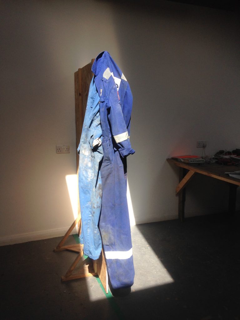 Two blue boiler suits - which were worn by Abel Shah on the residency - hang on top of each other in the SSW main studio, which has white walls and a mid grey coloured floor. Sun coming through a window in the roof casts a bright box of light into the studio, highlighting the boiler suits and putting the divider they hang on and the rest of the studio into darkness. A desk with some things on it is just about visible on the right-hand edge of the image.