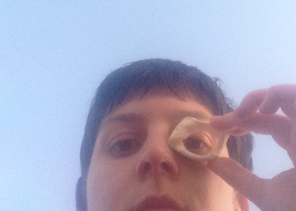 Selfie of Juliet’s face from the upper lip upwards. Juliet is a white person with brown hair in a mullet style and brown eyes. Juliet is gazing softly down at the camera and holding a broken cream coloured shell to their right eye which is looking through the shell as if it was a monocular. The image is soft in focus and the evening’s golden light is touching a side of the face, surrounded by light blue sky.