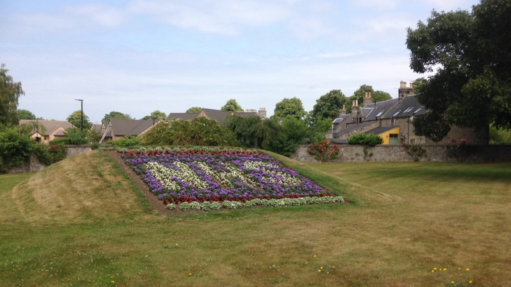 A flower bed planted on a grassy mound in Forres, a small town in Moray. White flowers fill out the letters ‘NHS’, the backdrop is formed of purple flowers with a trim of red flowers.The grass on the mound and the lawn the mound sits on is trimmed short and the grass looks to be yellowing from dryness. Behind the mound is a wall with some houses and trees on the other side.