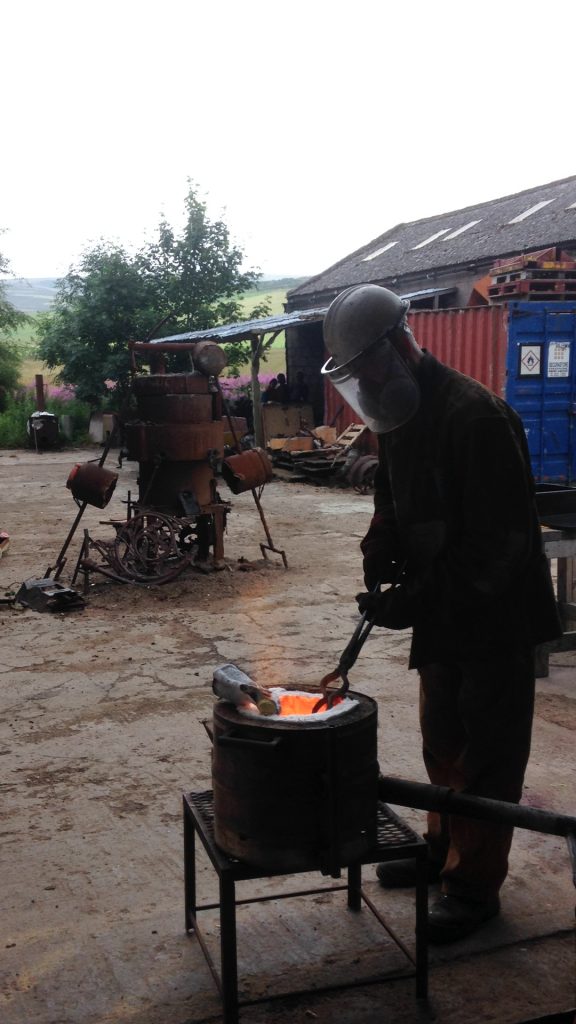 A person stands over a small furnace outside in the Scottish Sculpture Workshop yard, in front of the forge and iron cupolette furnace. The person has dark shadow cast over them. They are wearing a visored, white helmet and a respirator mask. They are looking into the furnace whilst using tongs to hold something in it. Whilst the furnace is also cast in shadow, a glowing orange colour is expanding from its interior.