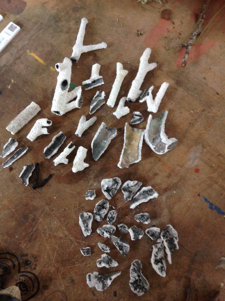Collection of bits of broken ceramic shell laid out on a brown studio table. The ceramic shell needs to be smashed once the bronze has been poured into it in order to retrieve the bronze casts. Around half of the pieces are from the twigs and are roughly between 2-10cm long. Their outside is white and their inside various shades of grey / light brown. The other half are from the parts of the shell that surrounded the lichen on the twigs.