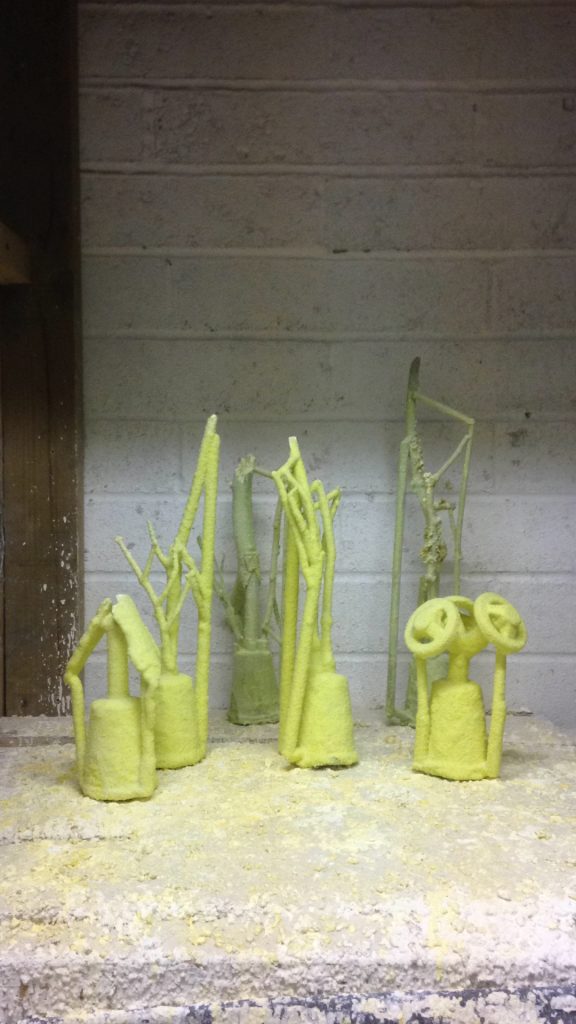 Six molds of twigs made from ceramic shell sit on a bumpy, textured surface waiting for their ceramic shell to dry. The molds are fluorescent lime green in colour. All six molds have a bell shaped base with thin cylindrical shapes coming off the base - these are called ‘runners’. The backdrop is a white breezeblock wall.