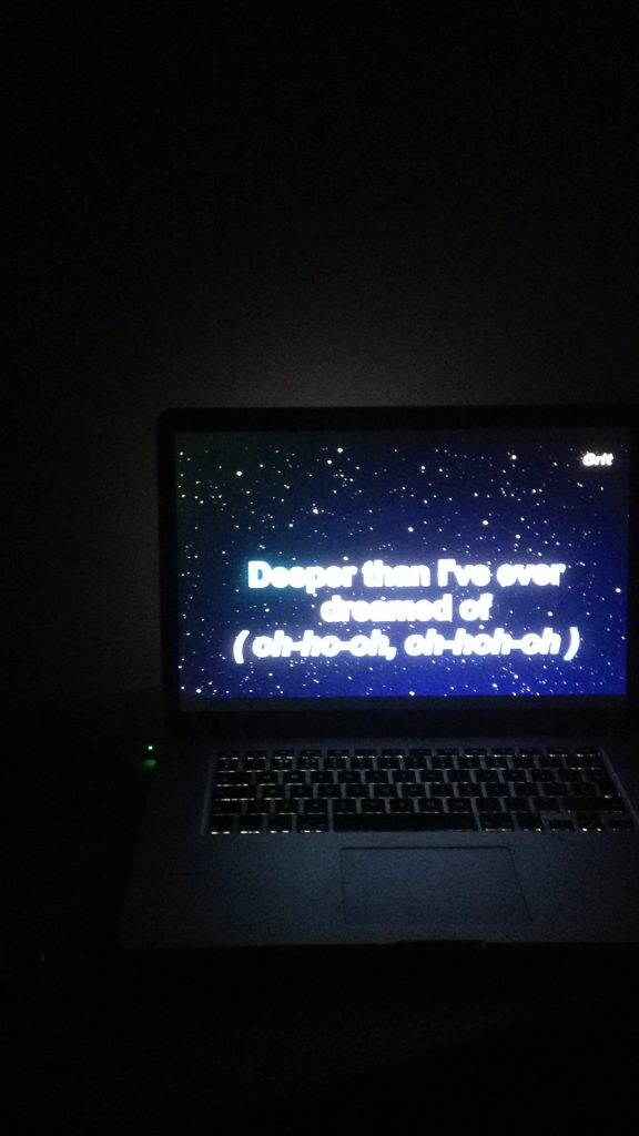 An image of a Macbook with the lyrics ‘Deeper than I’ve ever dreamed of (oh-ho-oh, oh-hoh-oh) in a white, thick, sans serif font. The lyrics sit on a dark blue, glitter starry background. They are from Maria Carey’s ‘Emotions’. The room is dark so the Macbook’s screen is strongly illuminated.