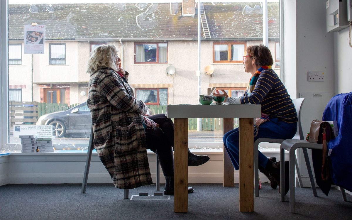 The profile of 2 people in a room, sat opposite each other at a table. The person on the left has blonde, wavy, shoulder-length hair. They wear a long coat with a tartan-like pattern, dark bottoms and boots. The person on the right has brown, straight hair in a bob. They wear red glasses; a multi coloured knit scarf; a purple and yellow striped top and blue trousers. Both people are smiling and looking out the large shop-front window next to them. The window faces on to a row of two storey; brown; pebble dashed; terraced houses on the Main Street in Lumsden.