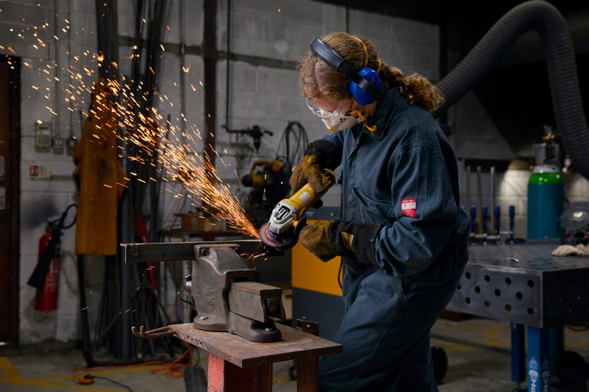 A person angle grinding in SSW's metal workshop. She is wearing a dark blue boilersuit and PPE. Sparks are flying from the grinder and metal.