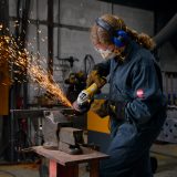 A person angle grinding in SSW's metal workshop. She is wearing a dark blue boilersuit and PPE. Sparks are flying from the grinder and metal.