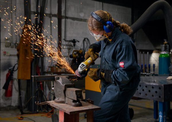 A person angle grinding in SSW's metal workshop. She is wearing a dark blue boilersuit and PPE. Sparks are flying from the grinder and metal.