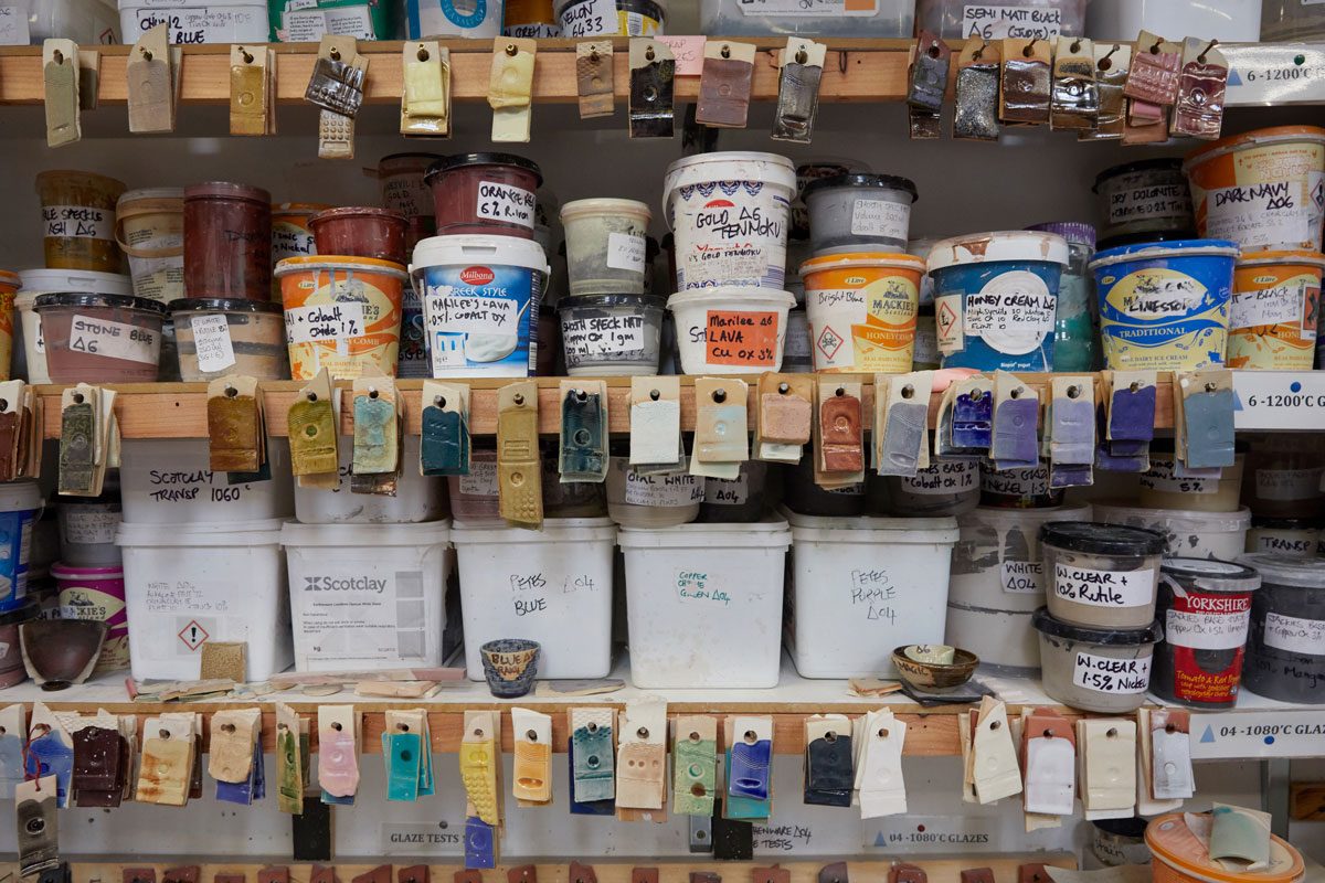 Three messy shelves on the wall. Stacked high with miscellaneous tubs containing glazes, labelled with handwritten stickers. Various test tiles hang from the front of the shelves in lots of colours and textures.