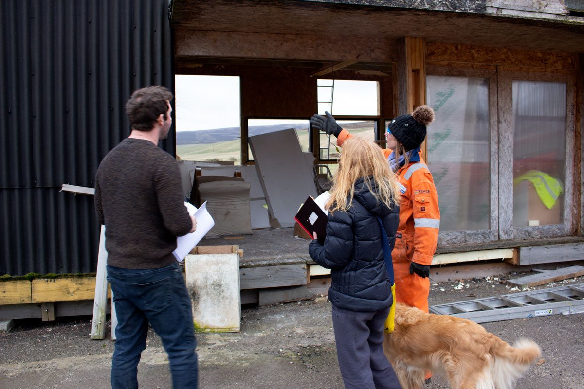 three people and a golden retriever dog stand in front of a chipboard building in a state of demolition. The person on the right, wearing an orange boiler suit and wooly hat gestures at the building. The other two are wearing jumpers, trousers and clutching papers and notebooks.