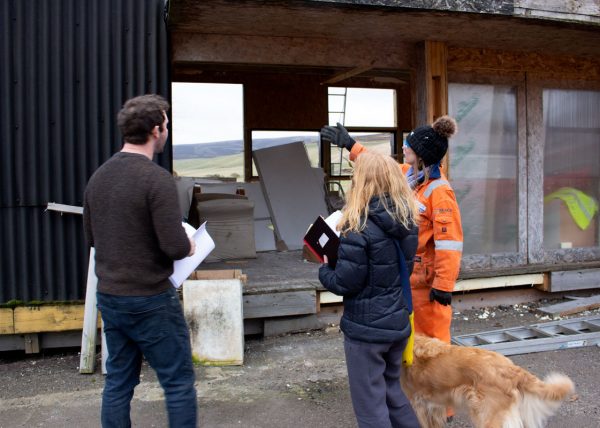 three people and a golden retriever dog stand in front of a chipboard building in a state of demolition. The person on the right, wearing an orange boiler suit and wooly hat gestures at the building. The other two are wearing jumpers, trousers and clutching papers and notebooks.