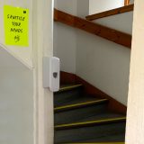 A fluorescent yellow paper sign with the words 'sanitise your hands' is on a white wall next to a staircase. The stairs curve up out of shot. There is a hand sanitiser pump on the entrance to the stairs.