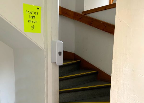 A fluorescent yellow paper sign with the words 'sanitise your hands' is on a white wall next to a staircase. The stairs curve up out of shot. There is a hand sanitiser pump on the entrance to the stairs.