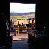 A view of the foundry looking out the large doors to the hills. The bright sunshine outside makes the inside very dark. Equipment and benches catch the light a little. Outside the iron furnace, forge and various parts sit in the yard.