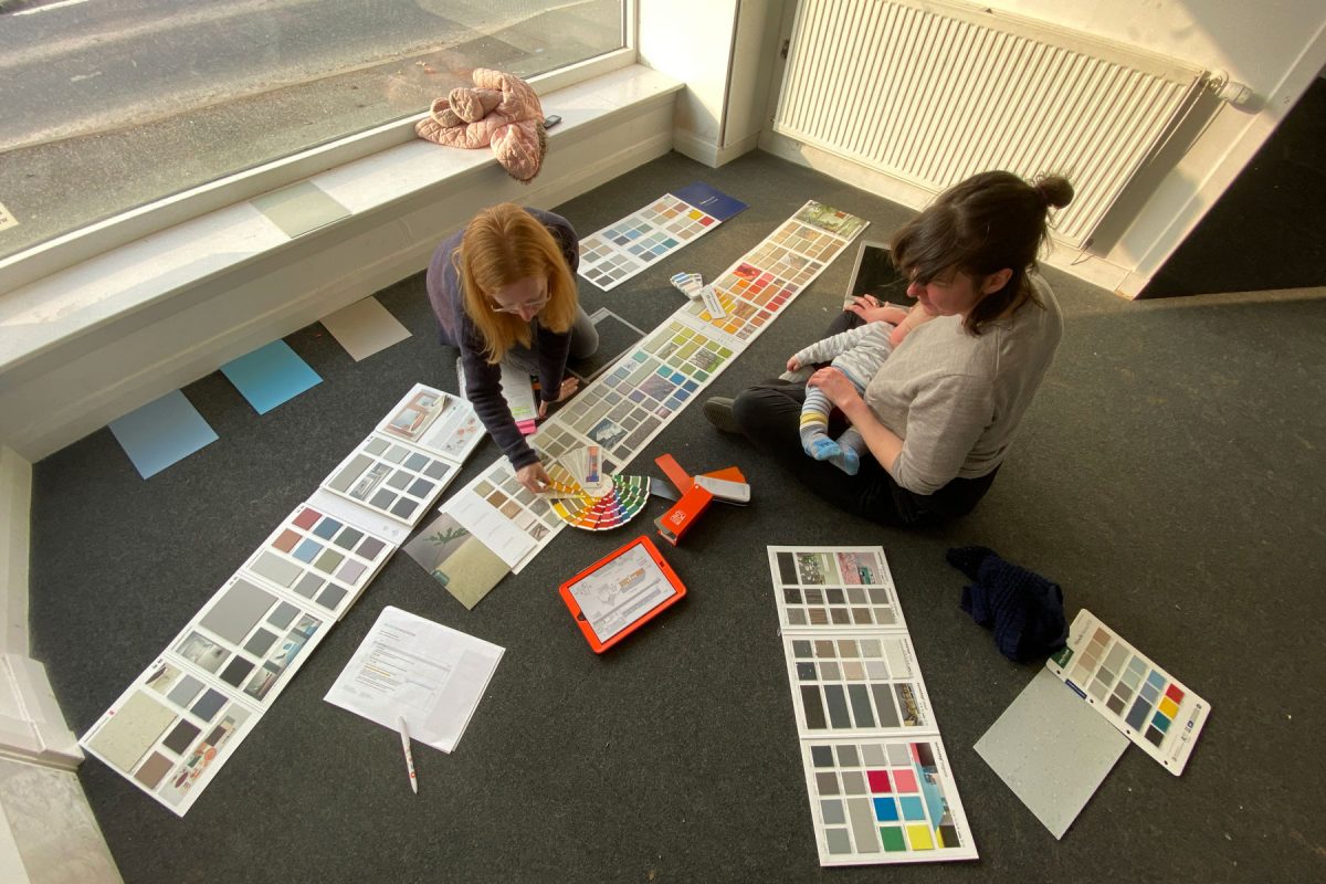 Two people sit on the grey carpet in the SSW old office. Colour samples are spread out on the floor around them and they are comparing different options. One person holds a baby in their lap.