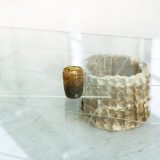 A photo of an artwork by Fionn Duffy. A small brown glass cup sits on top of a glass table top. The cup is made from seaweed ash and beach sand. There is a grid drawn with fish glue on the table top, and it is held up by a ceramic vessel which is also brown. Between the vessel and the glass table top is the corner of another sheet of glass, there is some writing etched into it, but it is hard to see what it says.