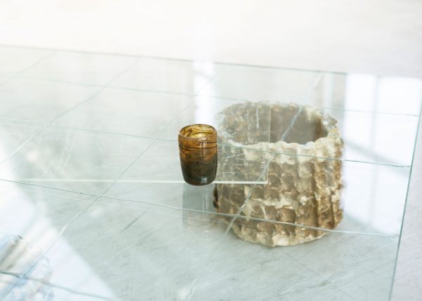 A photo of an artwork by Fionn Duffy. A small brown glass cup sits on top of a glass table top. The cup is made from seaweed ash and beach sand. There is a grid drawn with fish glue on the table top, and it is held up by a ceramic vessel which is also brown. Between the vessel and the glass table top is the corner of another sheet of glass, there is some writing etched into it, but it is hard to see what it says.