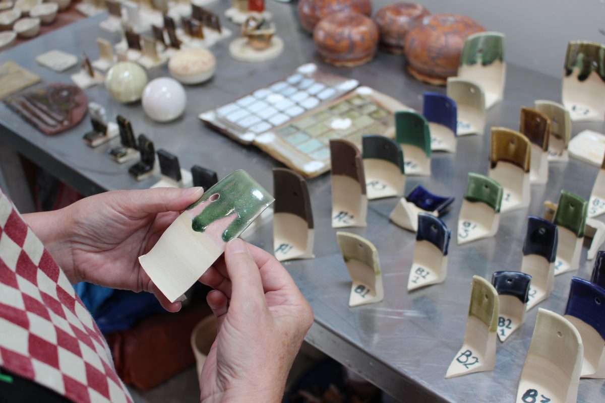 A person to the left of the image is holding a glaze test tile with an emerald green glaze at the top. Underneath there are a selection of glaze test tiles in a range of greens, blues and browns. The person holding the test tile is wearing a checked apron, only the apron and hands are visible.