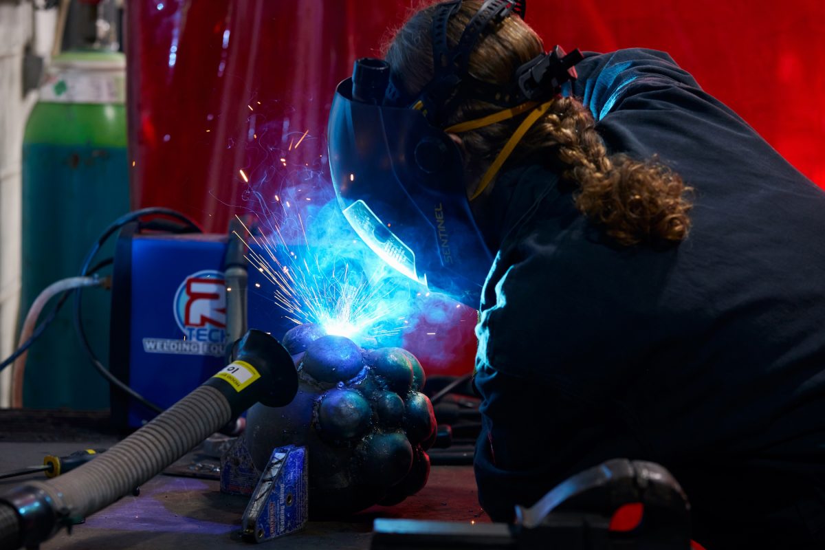 Artist Jodi Le Bigre is wearing a welding mask and joining metal spheres together in the SSW Metal Fabrication workshop. Her hair is in a plait and there is a red welding screen in the background. To the left of the image there is a welder and welding extraction.