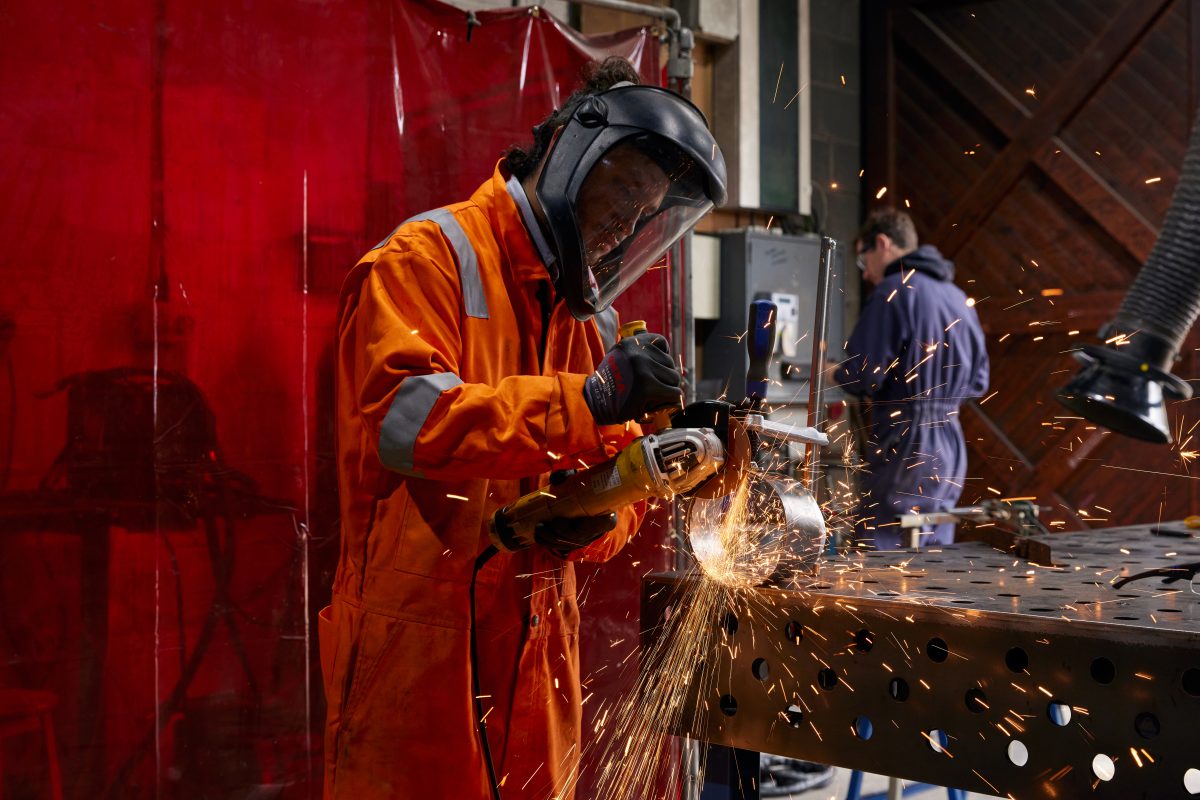 Image Description: Artist Dani Andres Ordoñez Muñoz working in the SSW Metal Fabrication workshop grinding a circular metal object with a red welding screen the the left of the image. Technical Director Ruaridh Allen is plasma cutting in the background to the right of the image.
