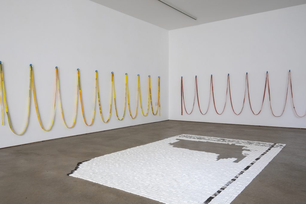 an exhitbition installation. A carpet of white tiles covers the floor and red and yellow ribbon loop along two walls.